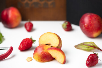 Ripe nectarines and fresh strawberries placed on a white table. In the background a red peeler and green leaves for salad or smoothie. Organic ingredients for making a healthy snack. Happy kitchen.