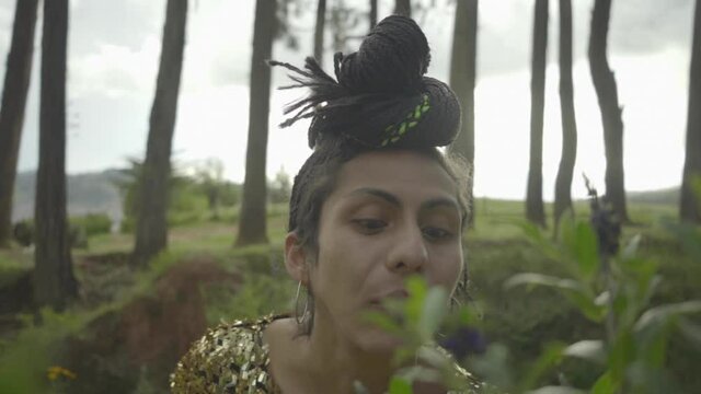Mujer transgénero con vestido dorado oliendo y jugando con plantas del bosque