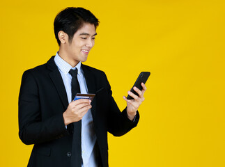 Man in formal black suit. Standing look at cellphone and smile seems get good news. Maybe about his credit card. Yellow background gives modern feel. The space can fill with text.