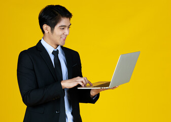 Young Asian businessman in a formal black suit. Standing work in hand holding laptop with smile. Seems he really enjoy his work. Yellow background gives a modern feel, empty space can enter text.