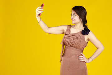 Middle aged women in brown dress and yellow backdrop. Stand, selfie of herself to send it to friends. Looks like take photo after make-up, before eating, it will become a fashionable now.