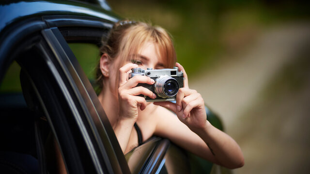 Young beautiful woman in car taking photos while leaning out the window