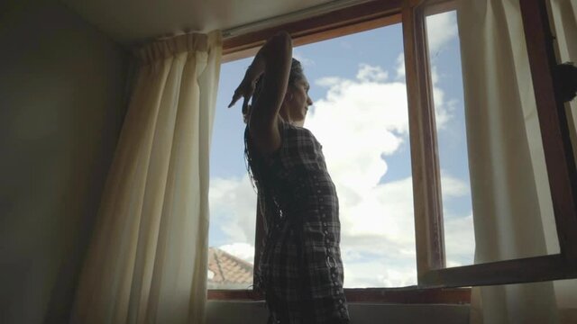 Mujer transgénero con trenzas arreglándose el cabello mientras observa el paisaje a través de su ventana 