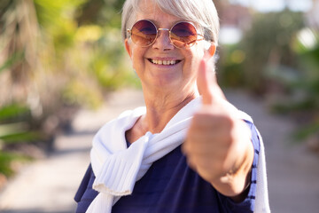 Portrait of attractive smiling senior woman in outdoors doing ok with hand. Defocused hand