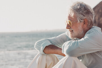Side view of handsome senior man, white haired, sitting on the beach looking at horizon over water