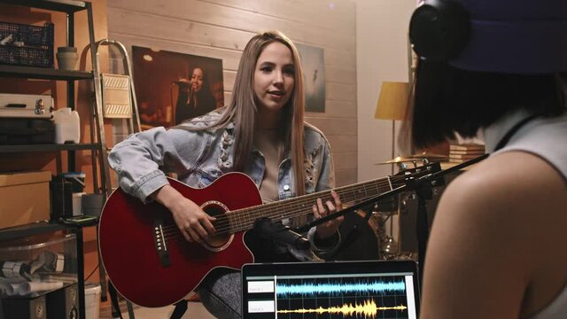 Over-the-shoulder medium slowmo of girl playing acoustic guitar in mic while recording music in retro-style music studio