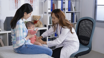 female doctor on swivel chair, gently pressing the baby girl's belly to check if she has abdomen problems. adorable toddler sitting in mother’s arms, getting routine checkups.
