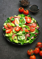 Fresh salad with salted salmon, avocado, egg, sesame seeds, olive oil, tomatoes and mixed herbs. Keto diet food. Black background, top view