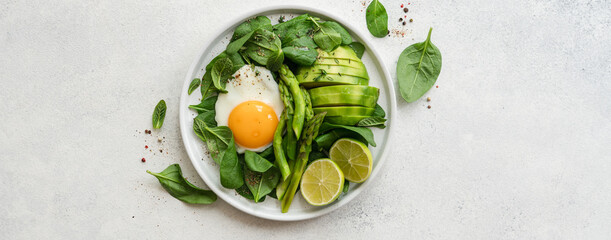 Healthy green breakfast with fried egg. Asparagus, spinach, avocado and egg in plate on white stone background