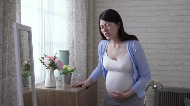 waist up japanese expectant mother propping herself on the cabinet is suffering labor contraction with a painful expression at home.