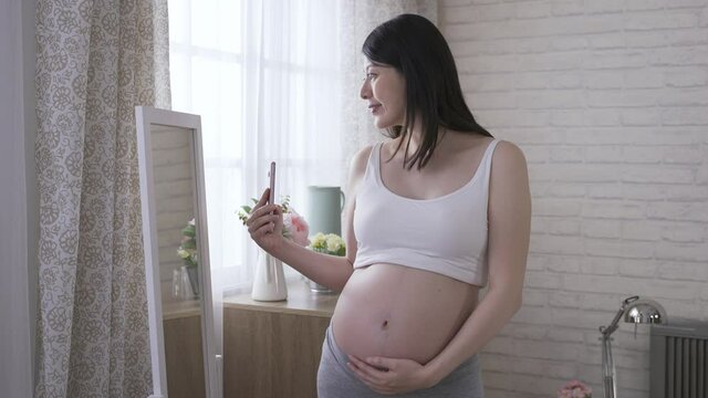 cheerful woman is tapping on the phone and smiling at her own reflection in the mirror while taking pictures during pregnancy at home.