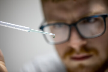 young bearded biologist in glasses stares intently at droplet of green chemical reagent on pipette....