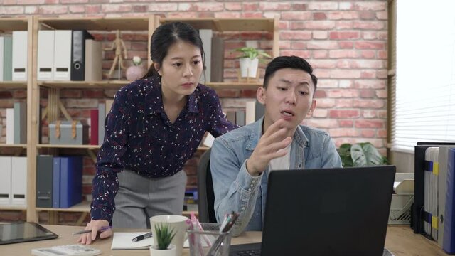 man sitting at office desk is talking and gesturing while discussing with his colleague who’s stooping over behind and looking at the monitor with him