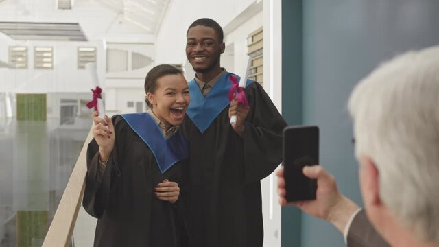 Back view of senior man making photo of young African student and his mixed-race female classmate wearing university graduate gowns using smartphone camera