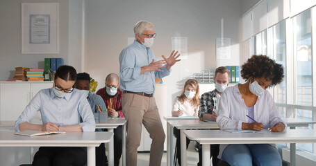 Mid adult professor wearing face mask while talking to a group of college students in lecture room