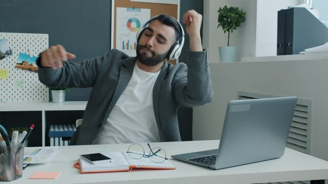 Funny businessman wearing headphones is dancing and working with laptop sitting at desk in office having fun alone at work. Youth and music concept.