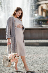 Happy pregnant woman with flowers walking near fountain.