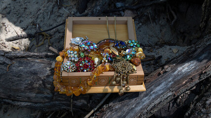 Open wooden box with glittering jewelry on the aged roots. Yellow Baltic amber necklaces, brooches with sparkling crystals and pearls. Luxurious texture, retro composition. 