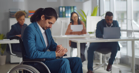 Sad disabled businessman sitting in wheelchair in office with colleagues working on background