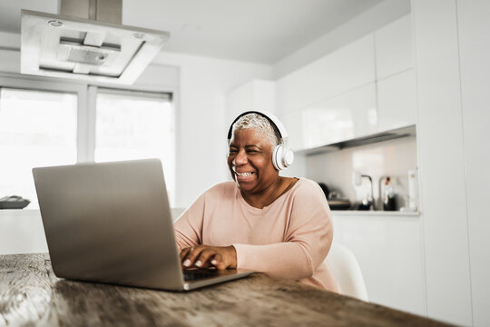 Senior african woman using laptop computer while wearing headphones at home - Focus on face