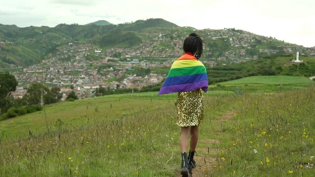 Mujer transgénero con vestido dorado y bandera LGTBQ caminando sobre la colina con vista a la ciudad de Cusco