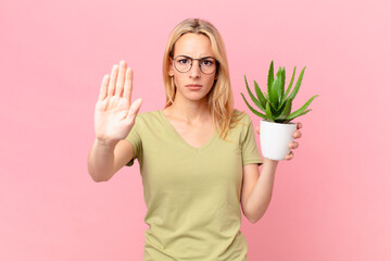 young blonde woman looking serious showing open palm making stop gesture and holding a cactus