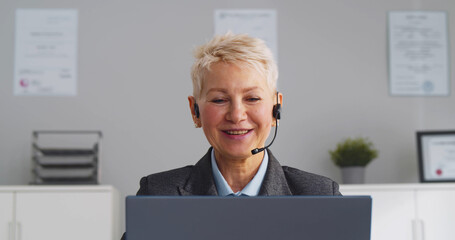 Mature businesswoman talking by headset while sitting in modern office