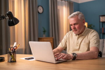 Elderly man sitting at desk with laptop in the evening. Senior works in a salon. Busy man in front of laptop screen. An elderly man shops online, pays bills, browses the news, checks email.