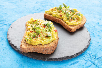 Two toasts with avocado, microgreens and hemp seeds on black slate board and on blue concrete background