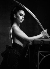 Young pretty brunette woman with naked upper body standing and holding dagger in hands over dark background. Martial arts and beautiful women concept