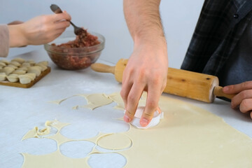 Man is cutting circles of dough for cooking dumplings using form, hands closeup. Woman is doing dumplings on table in kitchen. Process of cooking dumplings at home.