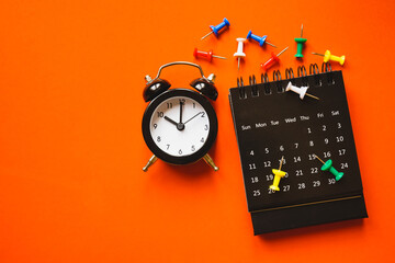 black analog alarm clock, opened calendar and multi color thumbtacks on grunge orange paper...