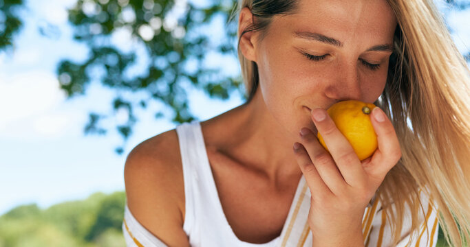 Closeup Of A Gorgeous Blonde Young Woman Enjoying Smells Of Lemon While Resting In The Park On A Sunny Day. Beautiful Female On A Picnic Having A Detox With Citrus Outdoors.