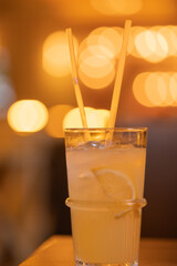 Lemonade in a glass with ice cubes on chopped lemon wedges. Refreshing cold drink with two straws in a cafe. Blurred warm background