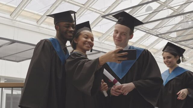 Medium shot of four diverse young women and men wearing university graduate gowns and hats standing together indoors, holding diplomas and making selfie using phone camera