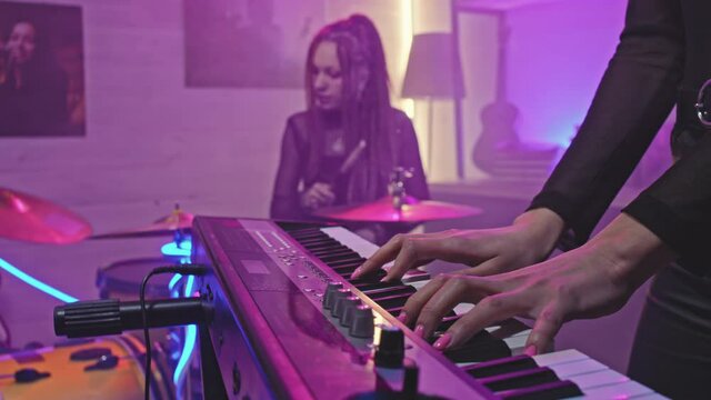 Slow-motion midsection closeup of unrecognizable female hands playing electronic keyboard synth during rock concert while female drummer with dreadlocks playing drums in background