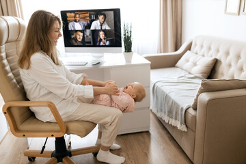 Obraz premium Stay at home mom working remotely on laptop, PC while playing and taking care of her baby. Young mother on maternity leave trying to freelance by the desk with infant child. Working mother concept