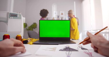 Hands of designer drawing sketches on paper in fashion design studio looking at laptop
