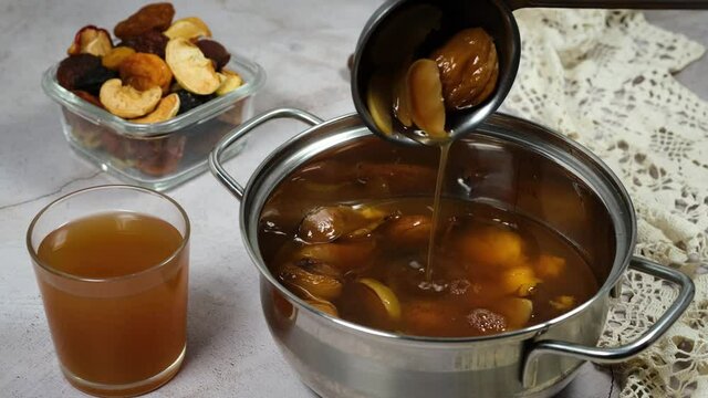Homemade compote making from dried fruits apples, apricots, plums etc. Person stirs fresh cooked vegetarian drink uzvar in saucepan with scoop.