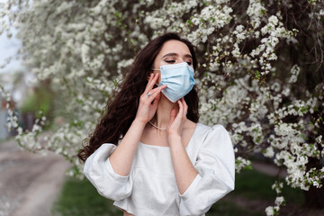 Girl in medical mask near white blooming trees in the park. Outdoor walking countryside at quarantine coronavirus covid-19 period. Spring lifestyle.