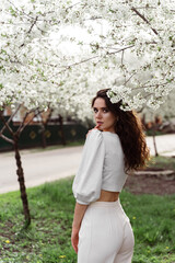 Spring lifestyle. Model posing near white blooming trees without mask outdoor countryside. Dreaming girl with curly hair.