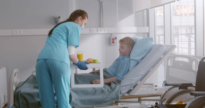 Nurse In Safety Mask And Gloves Serving Child Patient Meal In Hospital Bed