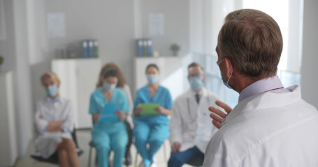 Obraz premium Mature doctor wearing white coat and safety mask giving presentation to healthcare specialists