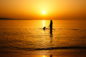 Tranquil scene with the silhouette of a woman and her dog having bathe in the sea during beautiful summer sunrise