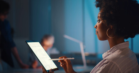 Professional afro-american architect studying blueprint on digital tablet in office working late