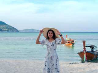 woman in dress holding wide brim hat on head standing at sea view sand beach, blue sea and sky on...