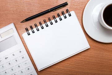 On a wooden table, there is a white cup of coffee, a white calculator, a pen and a white blank notebook with a place to insert text. Template. Business concept