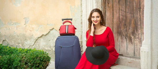 A young caucasian woman is sitting on the threshold of an old house in a red long dress with...