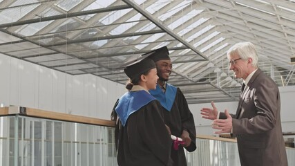 Medium shot of aged male university tutor talking to two African graduates wearing university graduate gowns and hats then shaking their hands and going away