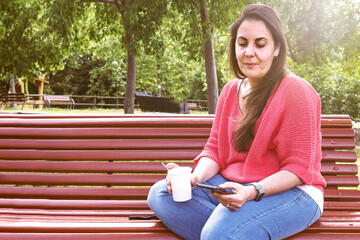 Woman holding a cup of cofee and a mobile device sitting outdoors at the park.